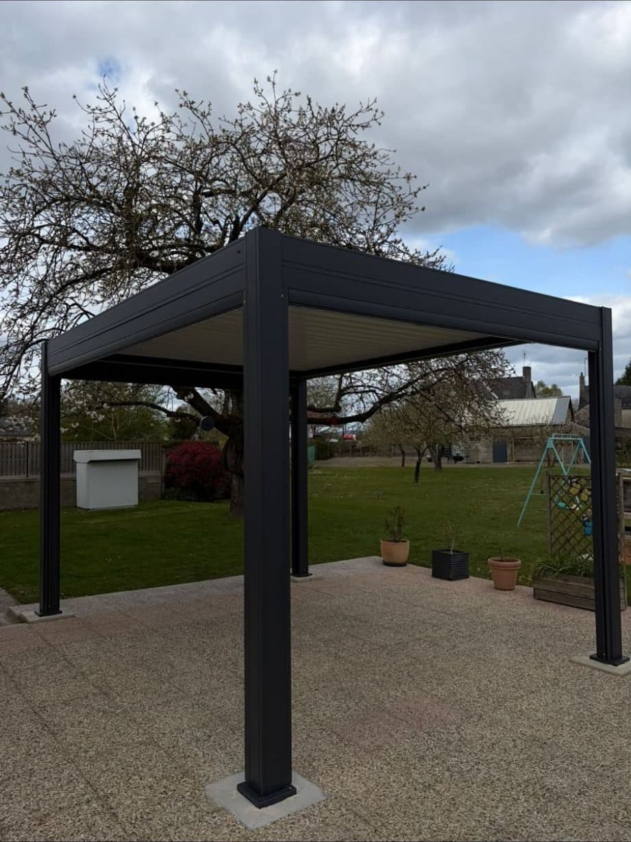 Pergola bioclimatique autoportante anthracite sur 4 poteaux, posée sur une terrasse au milieu d'un jardin