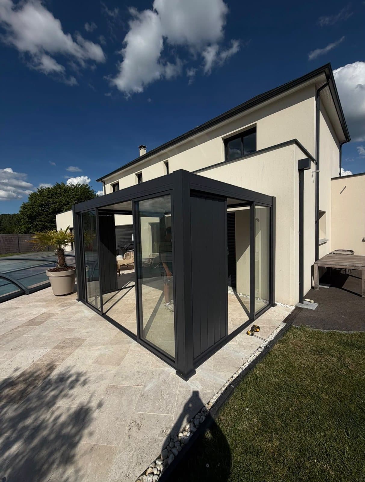Pergola bioclimatique anthracite avec parois vitrées et panneaux pleins, sur une terrasse en pierre près d'une piscine
