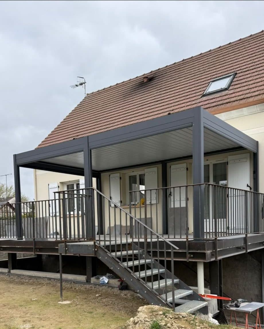Pergola bioclimatique autoportante posée sur une terrasse surélevée en métal avec escalier, devant une maison
