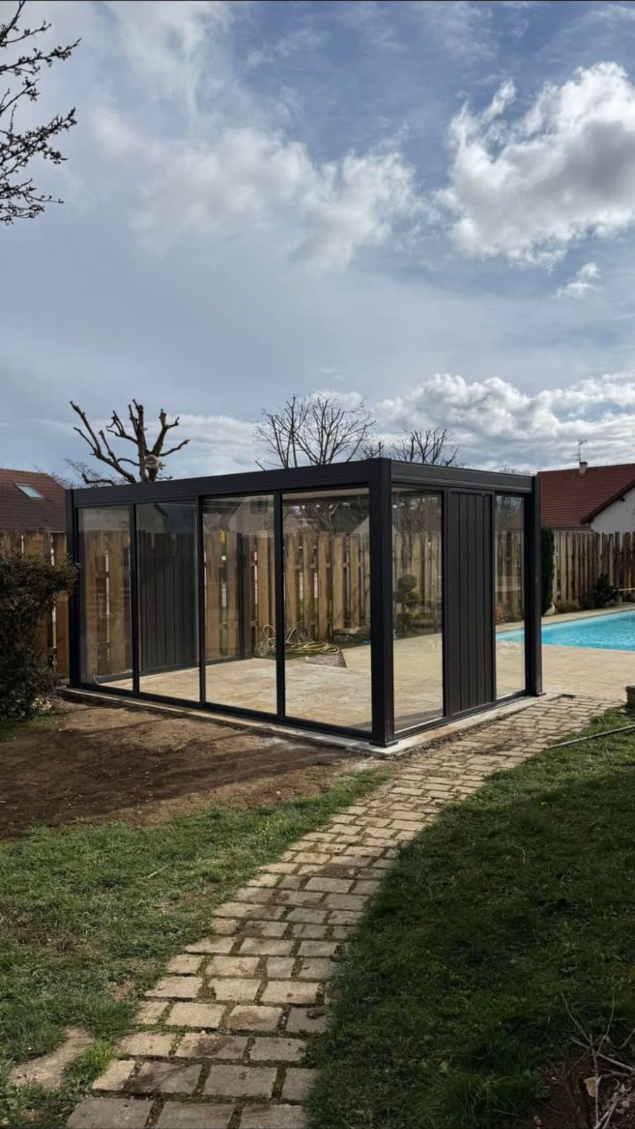 Pergola bioclimatique vitrée anthracite installée au bord d'une piscine, dans un jardin clos