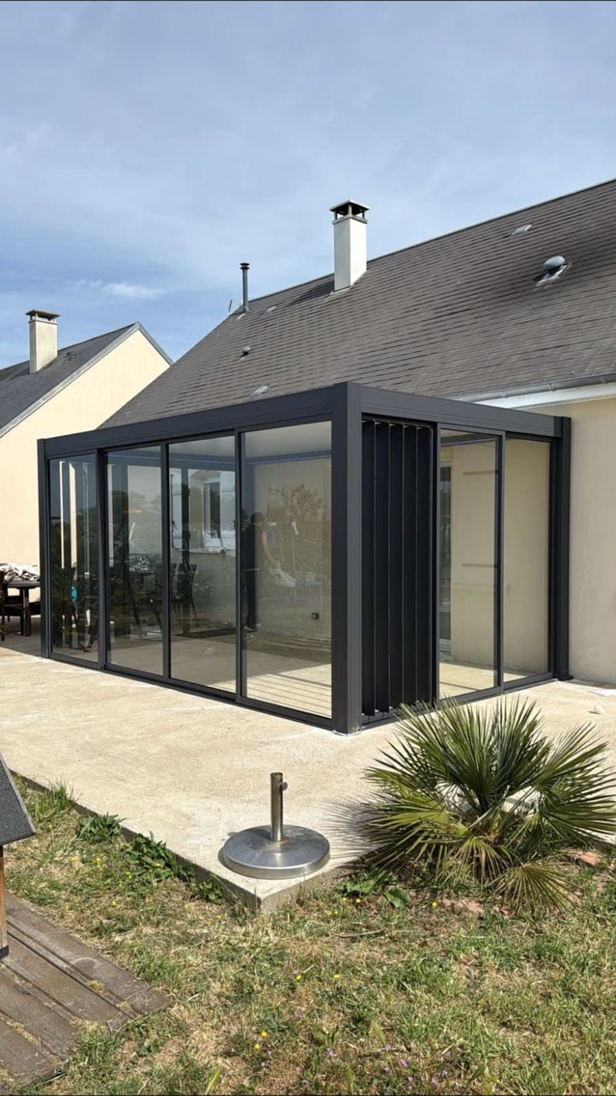 Pergola bioclimatique vitrée anthracite avec lames verticales orientables sur le côté, sur une terrasse en béton