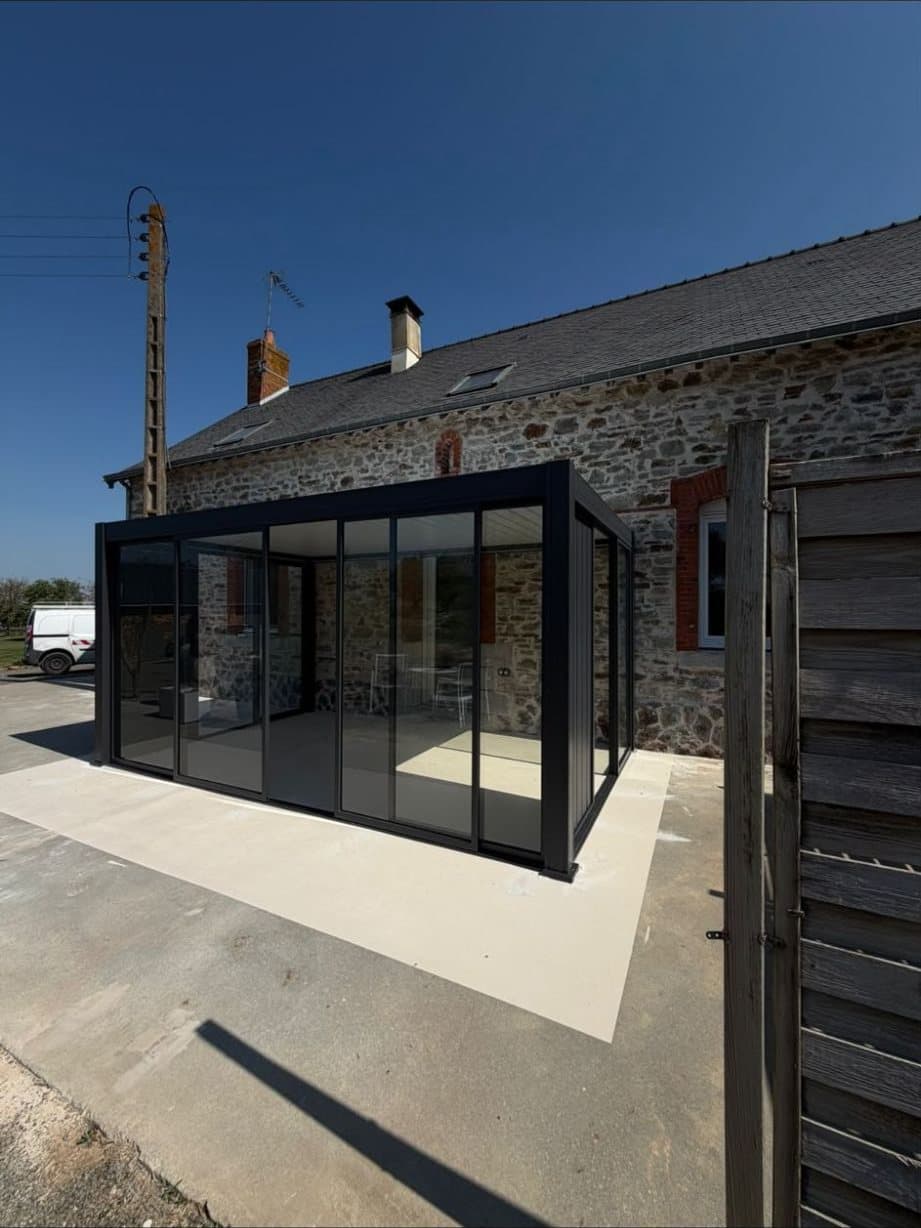 Pergola bioclimatique vitrée anthracite installée devant une longère en pierre, sous un ciel bleu