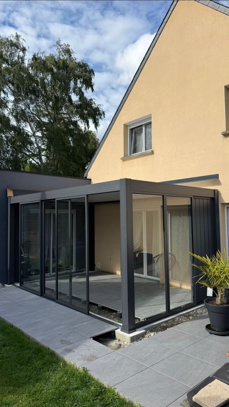 Pergola bioclimatique fermée par des vitrages coulissants, sur une terrasse en carrelage gris devant une maison