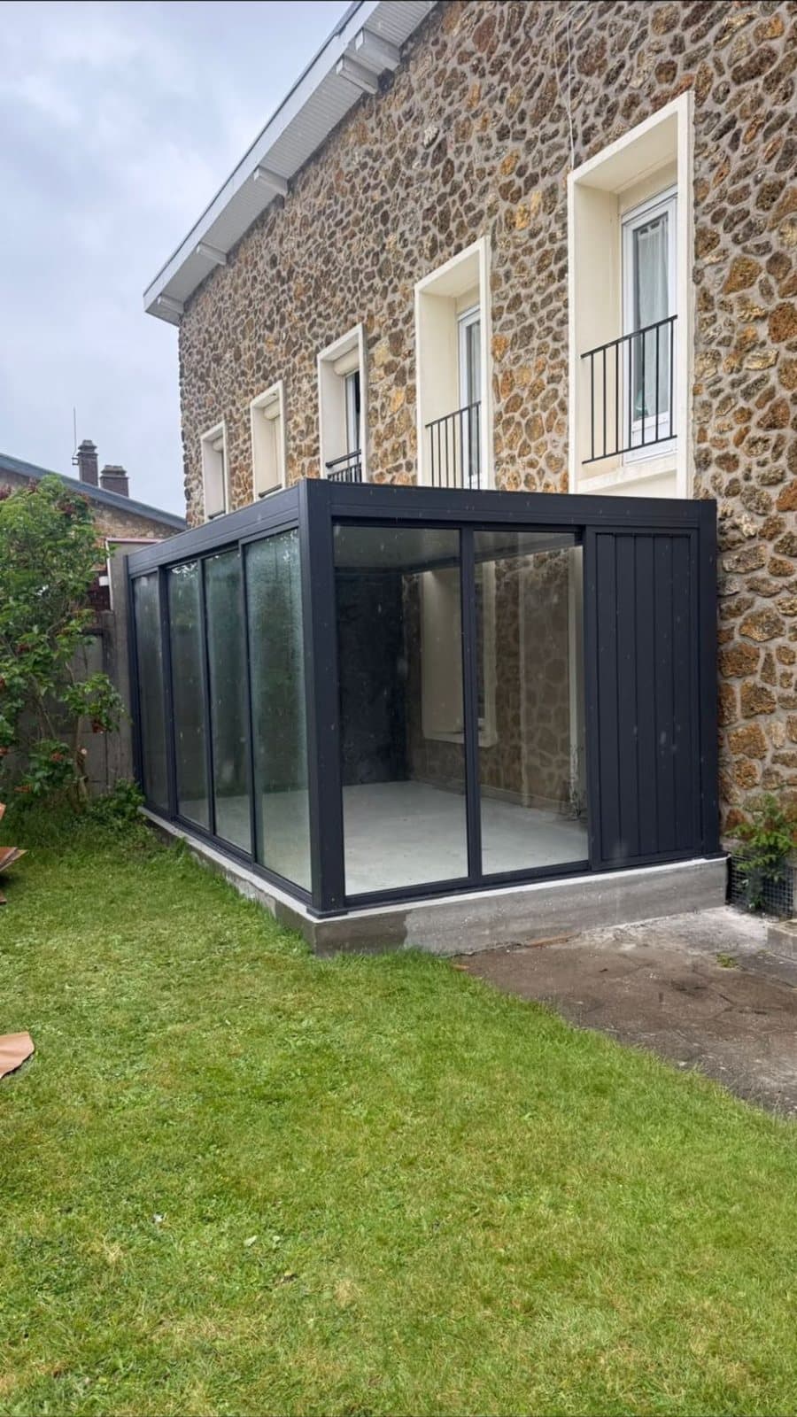 Pergola bioclimatique vitrée anthracite installée dans le jardin d'une maison en pierre meulière