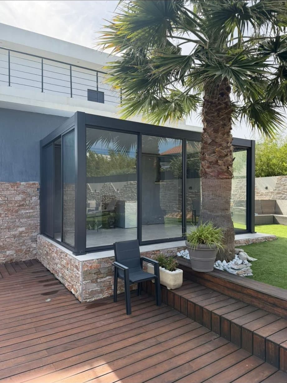 Pergola bioclimatique vitrée anthracite sur un muret en pierre, à côté d'un palmier et d'une terrasse en bois