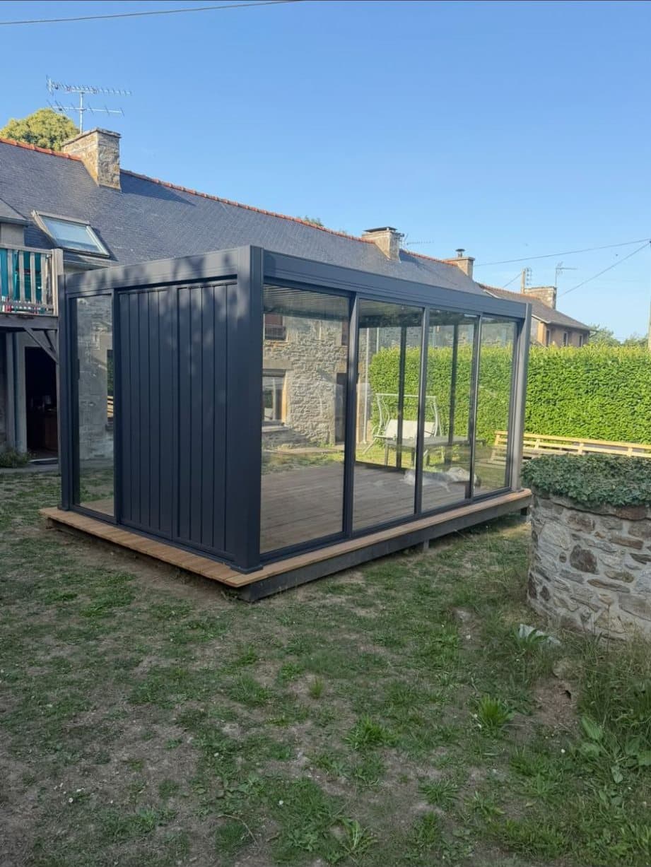 Pergola bioclimatique avec vitrages coulissants posée sur une terrasse en bois, dans le jardin d'une maison en pierre