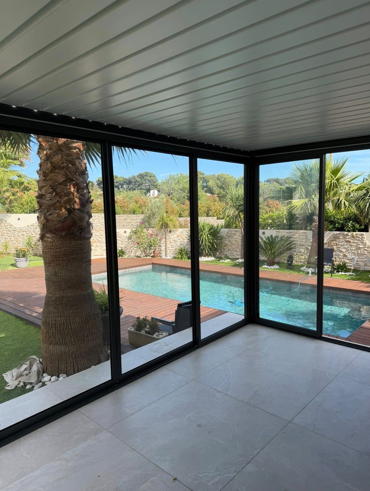 Vue depuis l'intérieur d'une pergola bioclimatique fermée par des vitrages, donnant sur une piscine et un jardin de palmiers